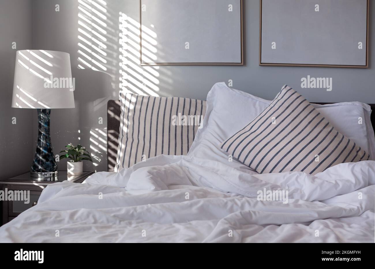 Early morning light casts light and shadows from window blinds onto striped accent pillows and blue and white covers on a bed Stock Photo