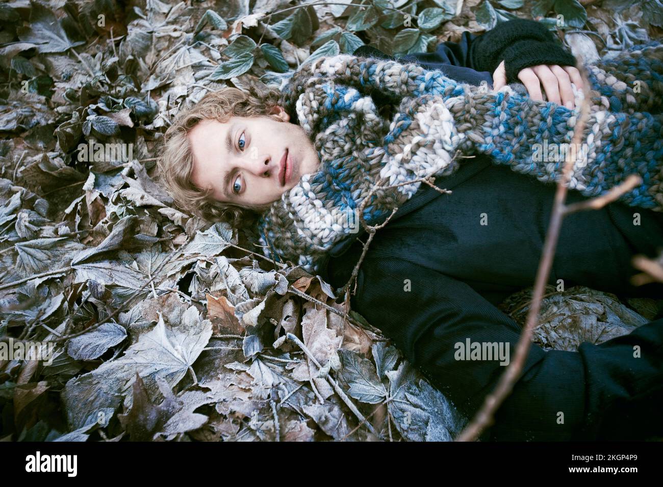 Young man dressed with a thick scarf laying down between icy leaves Stock Photo