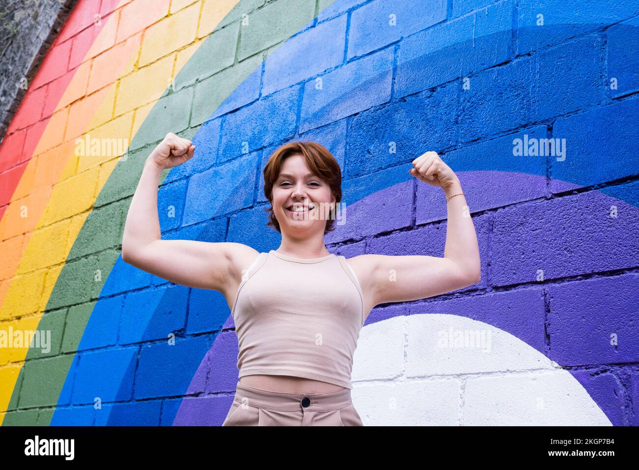 Happy young woman flexing muscles in front of rainbow flag painted on wall Stock Photo
