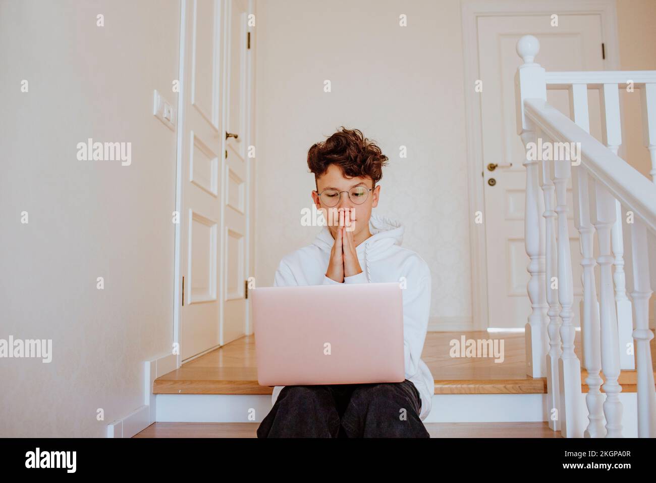 Boy with hands clasped sitting with laptop on stairs at home Stock Photo