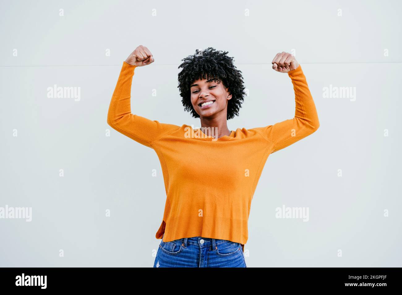Smiling Afro woman gesturing strength in front of white wall Stock ...