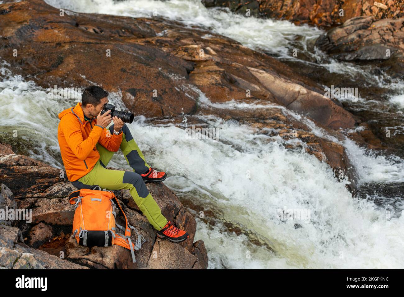 Mature man taking photos through camera sitting on rock by river Stock Photo