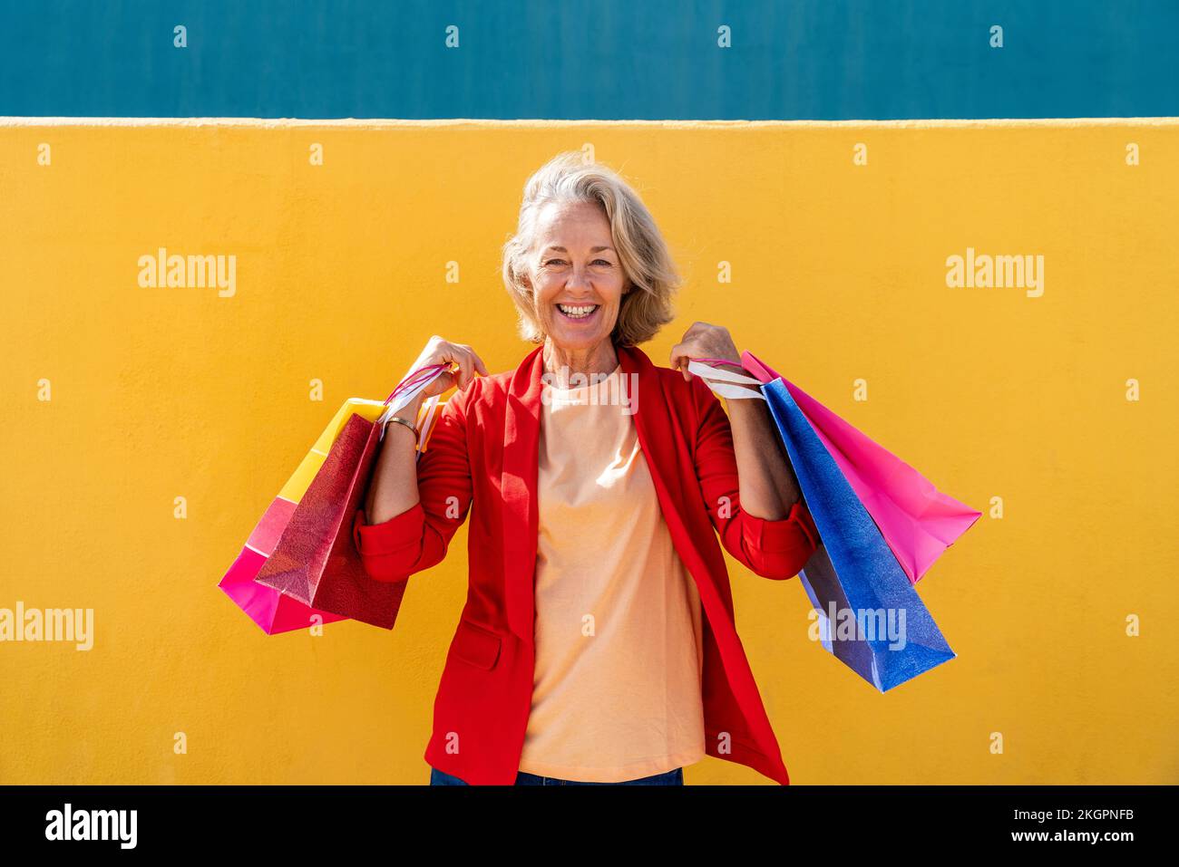 Happy mature woman with multi colored shopping bags in front of yellow wall Stock Photo