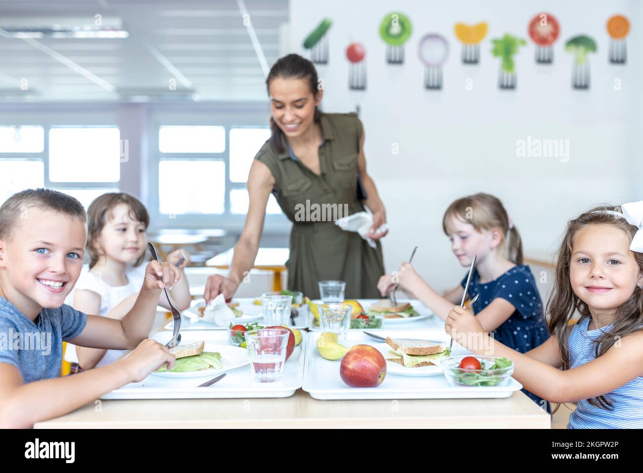 Smiling school students having lunch together with teacher at cafeteria