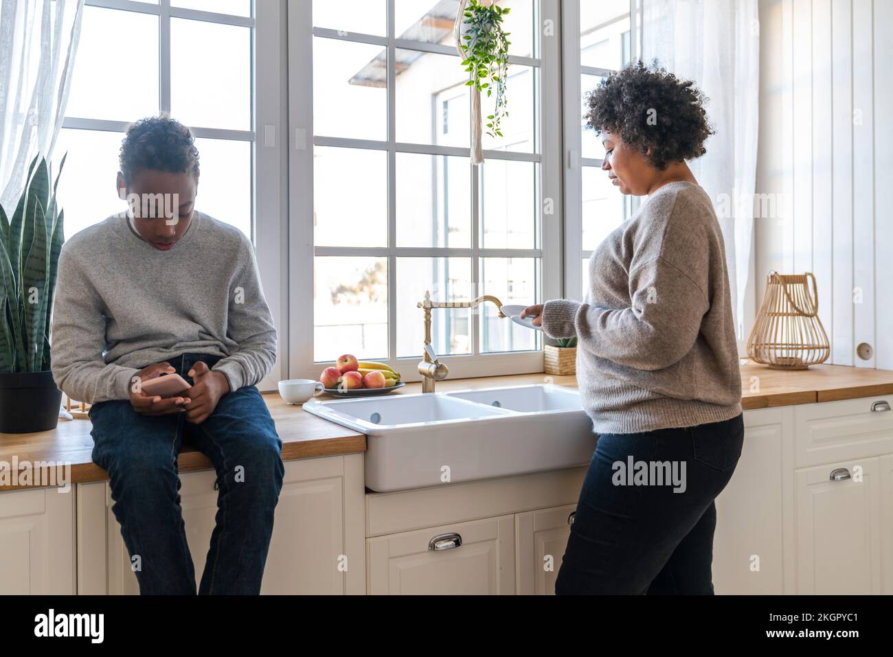 Son using mobile phone by mother washing utensils in kitchen Stock Photo