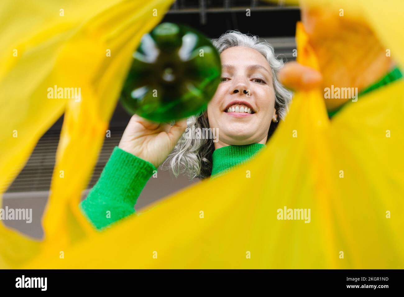 Smiling woman putting green bottle in yellow plastic bag Stock Photo