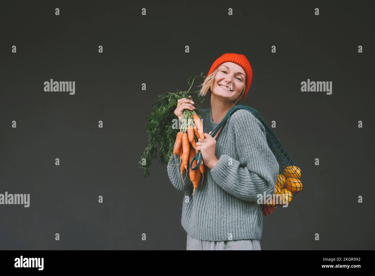 Happy woman holding carrots and mesh bag in front of gray wall Stock Photo