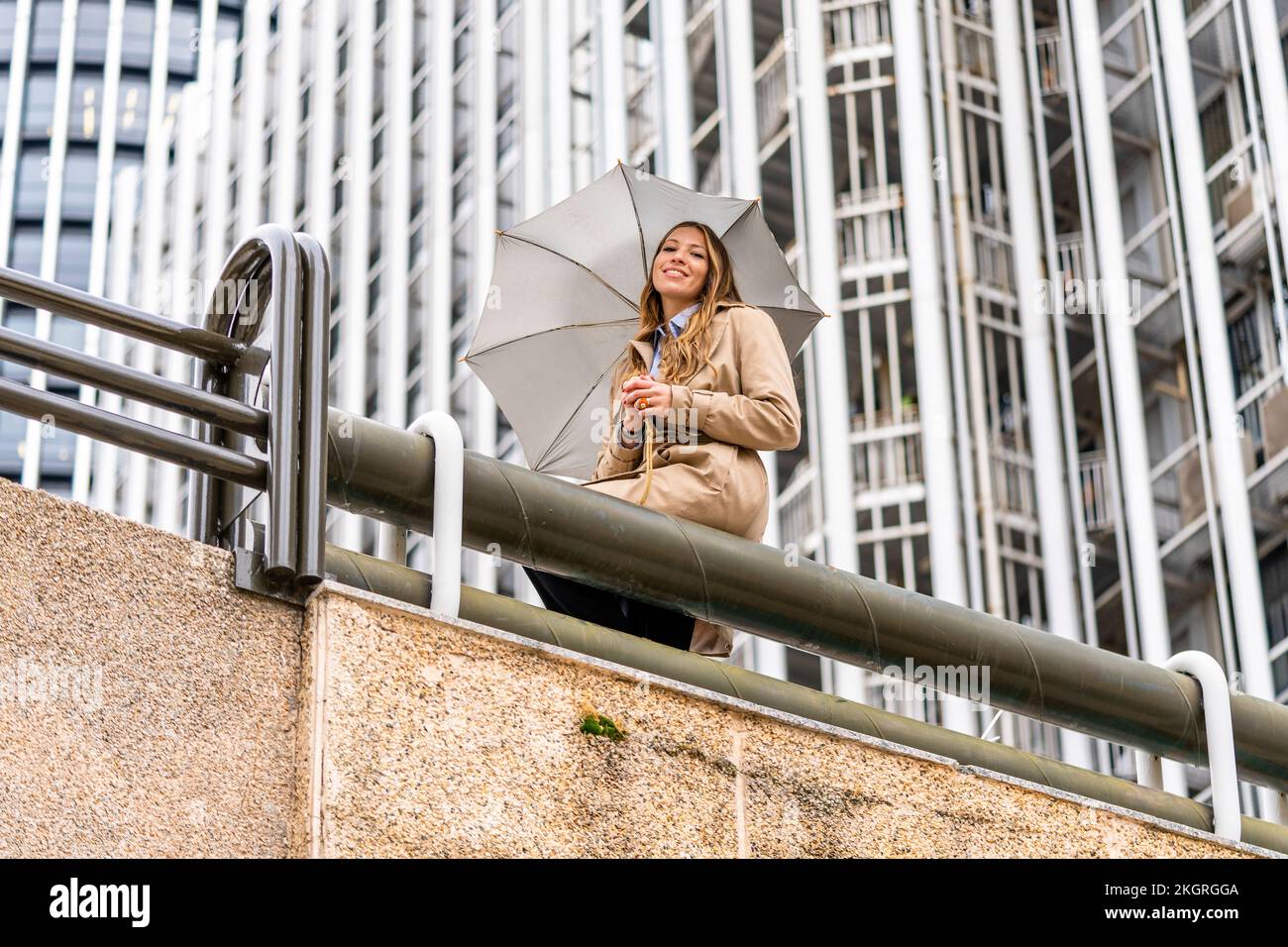 Smiling businesswoman holding umbrella sitting on railing in front of office building Stock Photo