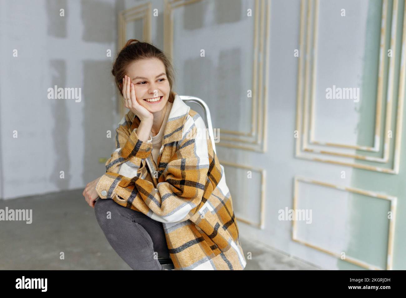 Smiling young woman in plaid shirt day dreaming with hand on chin in apartment Stock Photo