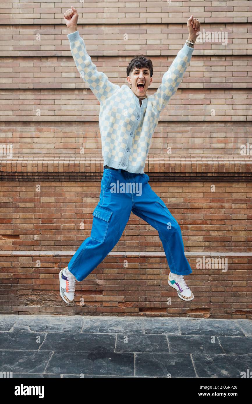 Happy young man jumping on footpath in front of brick wall Stock Photo