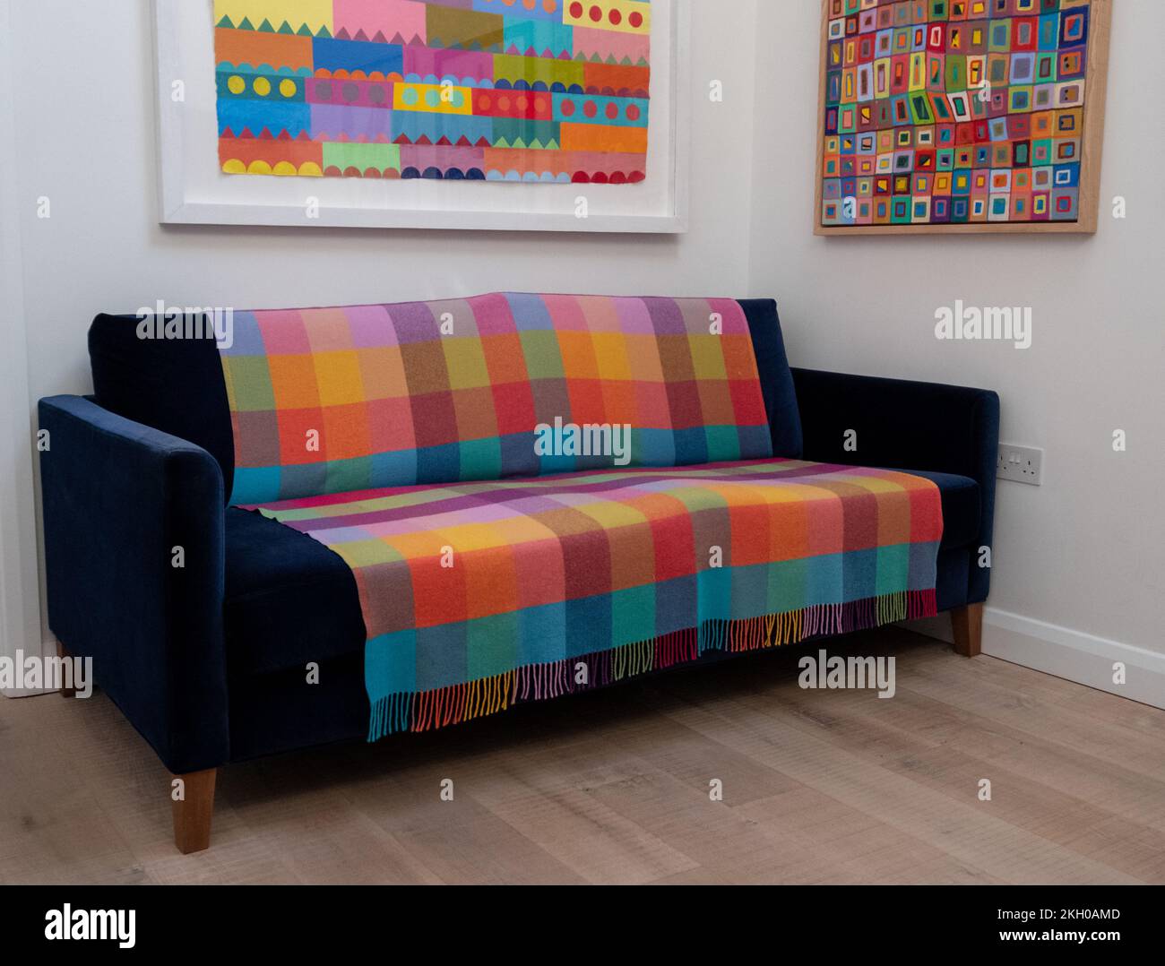 Blue velvet sofa with colourful wool throw blanket, in minimalist setting with white walls, oak