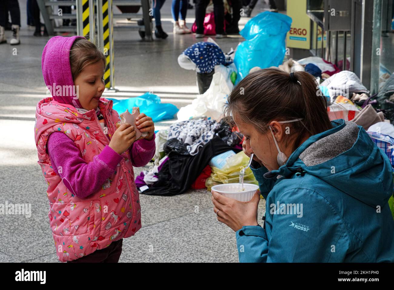 A woman sits squatting on the floor of a hall at Munich Central Station. She has a plastic cup in her hand containing soup. Her daughter is eating. Stock Photo