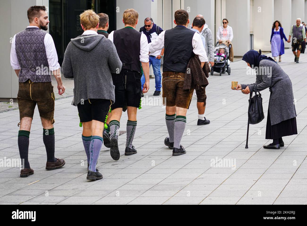 Young lads in Bavarian dress on their way to the Oktoberfest pass a beggar woman asking for money. Stock Photo