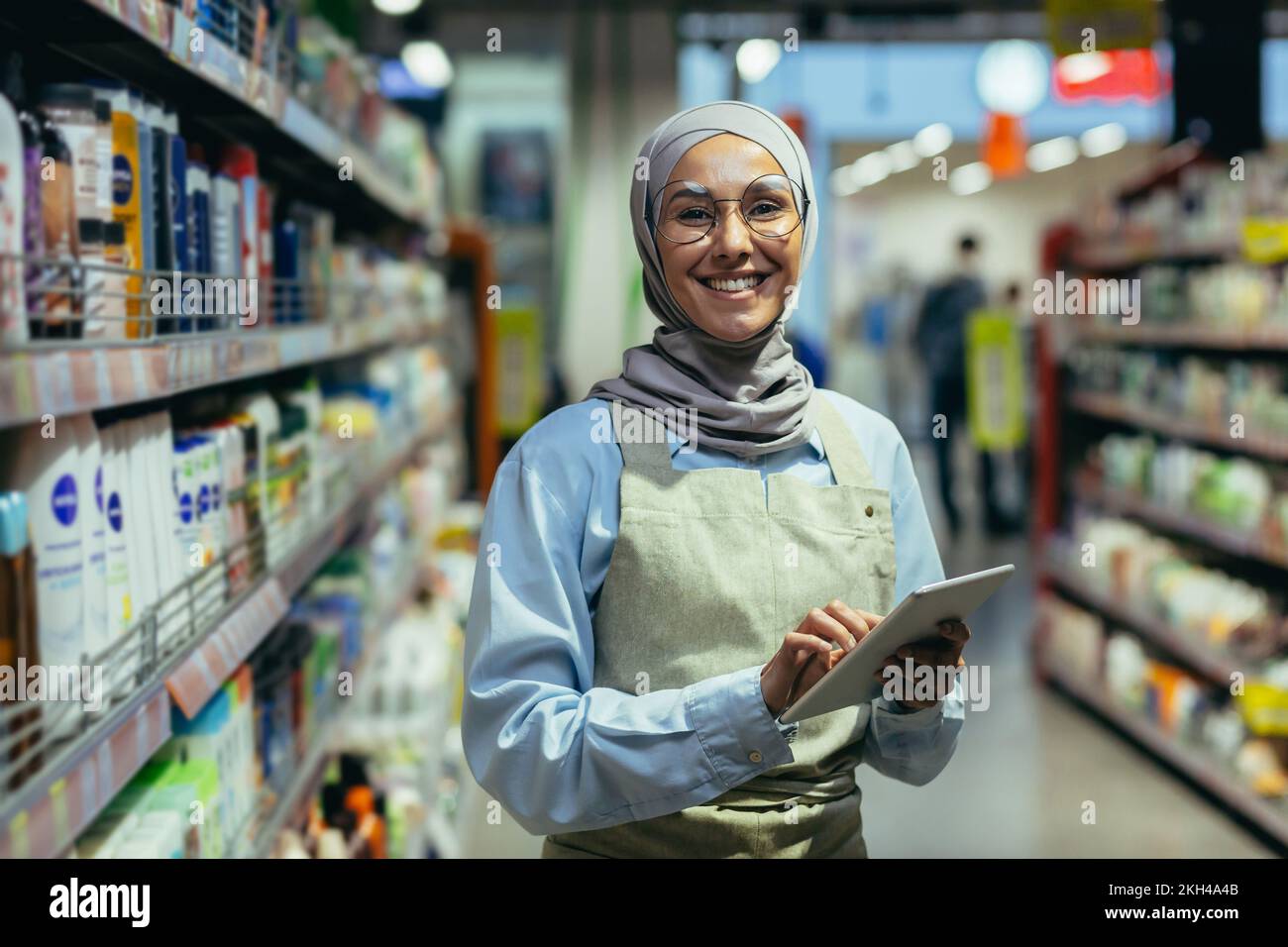 Portrait of a woman in a hijab, a shop worker in a supermarket with a tablet computer, looks at the camera and smiles, a female seller consultant in glasses among rows of shelves with goods. Stock Photo