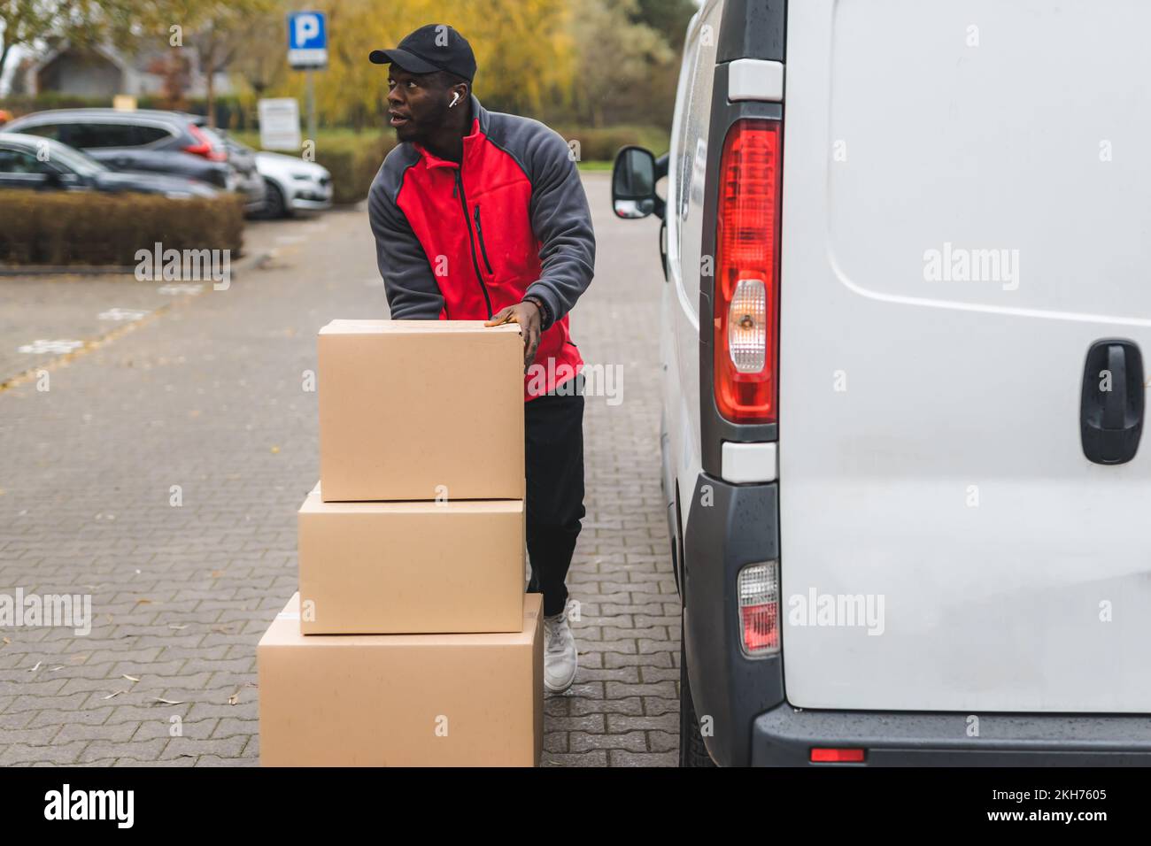 Young adult black delivery man standing next to white van with unloaded parcels stacked on trolley. Horizontal outdoor shot. High quality photo Stock Photo