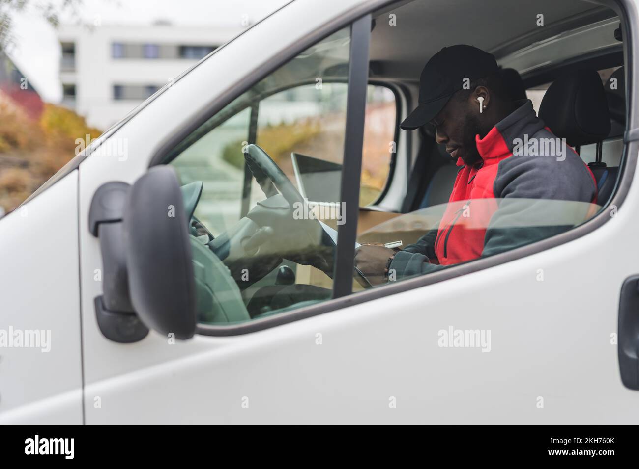 Technology used in package delivery process. Outdoor view of professional focused African-American post delivery man sitting in white delivery truck on driver's seat and looking at his check-list with white wireless earphone in his ear. High quality photo Stock Photo