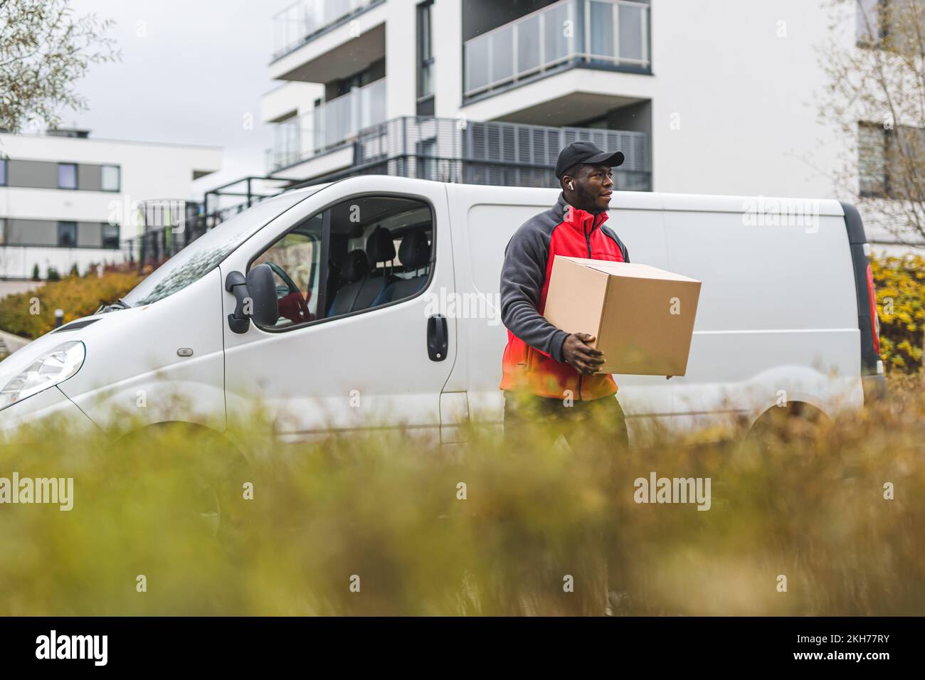 Black blue-collar worker - package delivery person - in front of white truck with cardboard package. Clothing - red jacket with gray sleeves and black hat. Blurred tall grass in the foreground. High quality photo Stock Photo