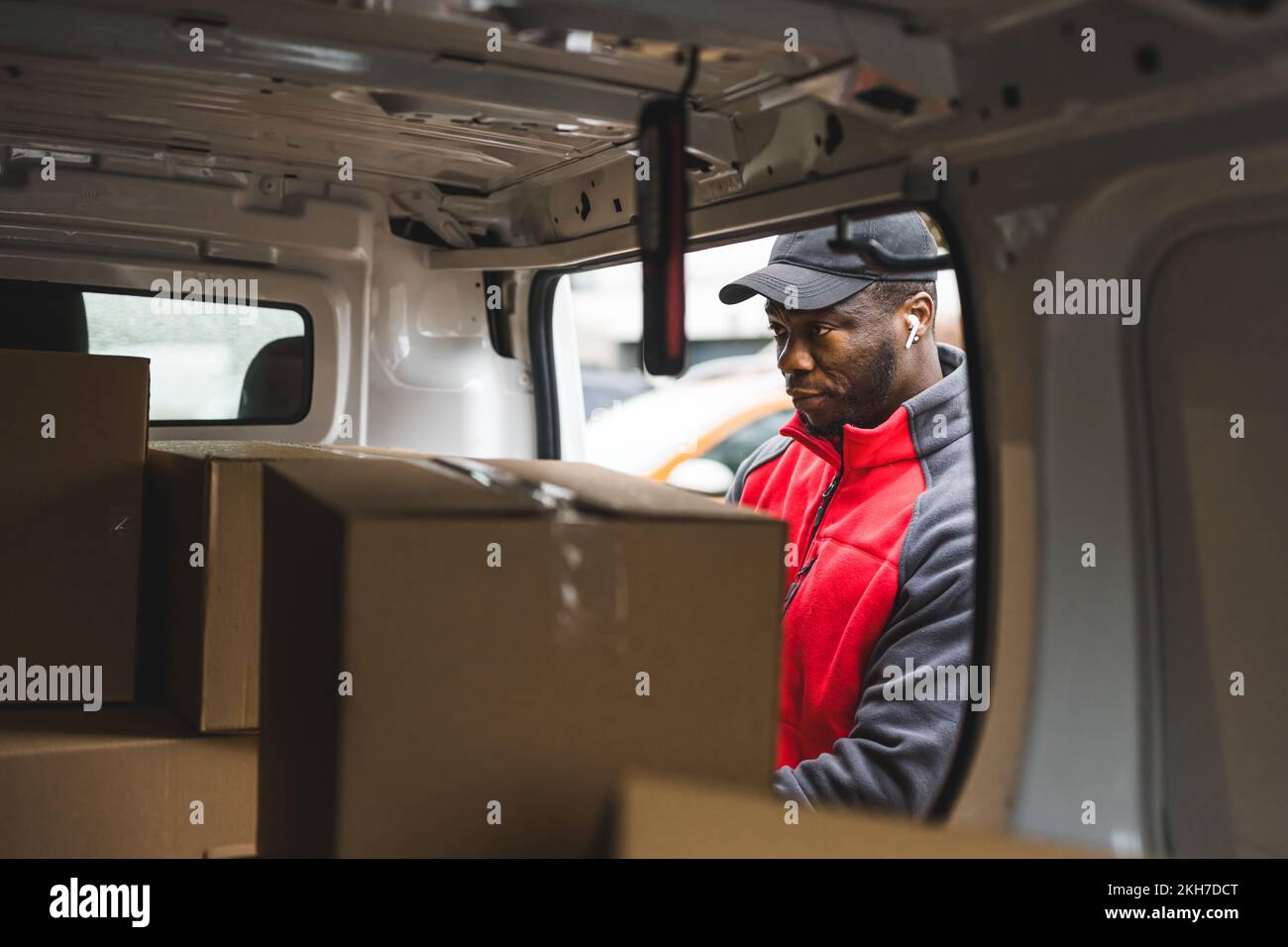 Young adult black delivery man wearing red uniform pullover looking inside van full of carboard box parcels. Horizontal shot. High quality photo Stock Photo