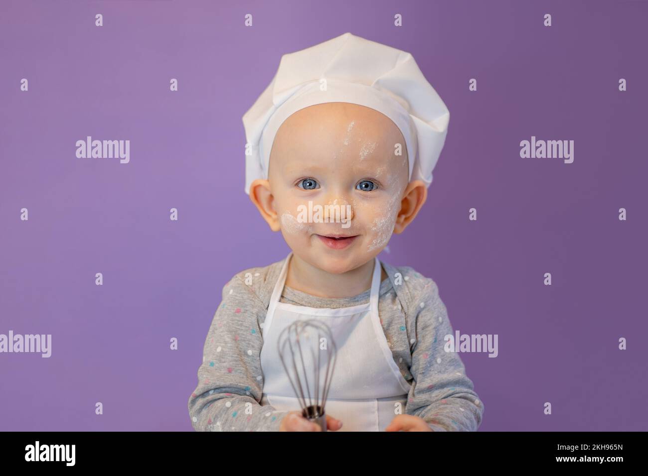 little cute baby in an apron and a chef's hat smiles on a lilac