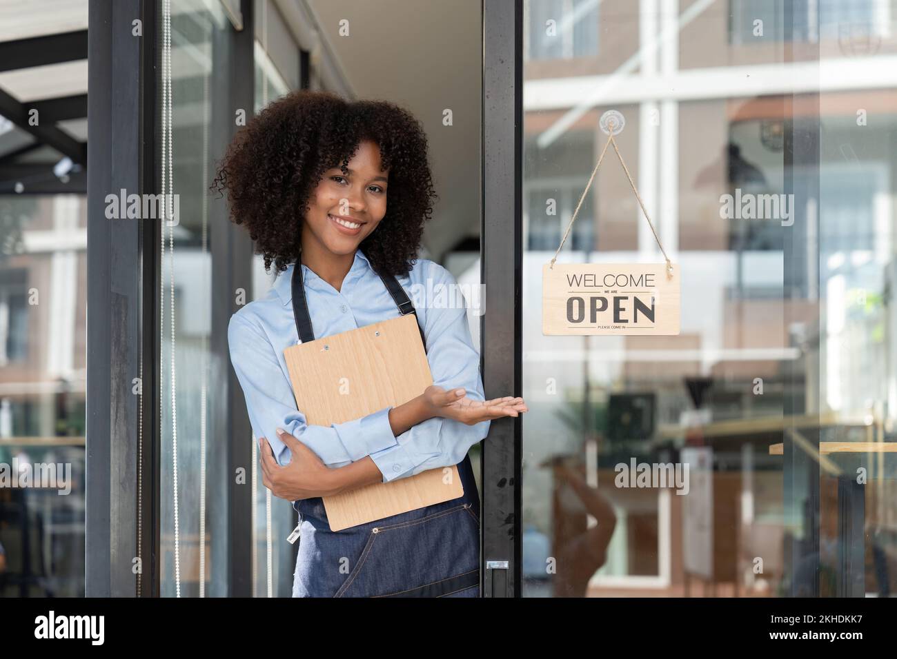 Small business african female owner smiling while turning sign for opening of cafe. Happy afro-american waitress entrepreneur in apron present sign on Stock Photo