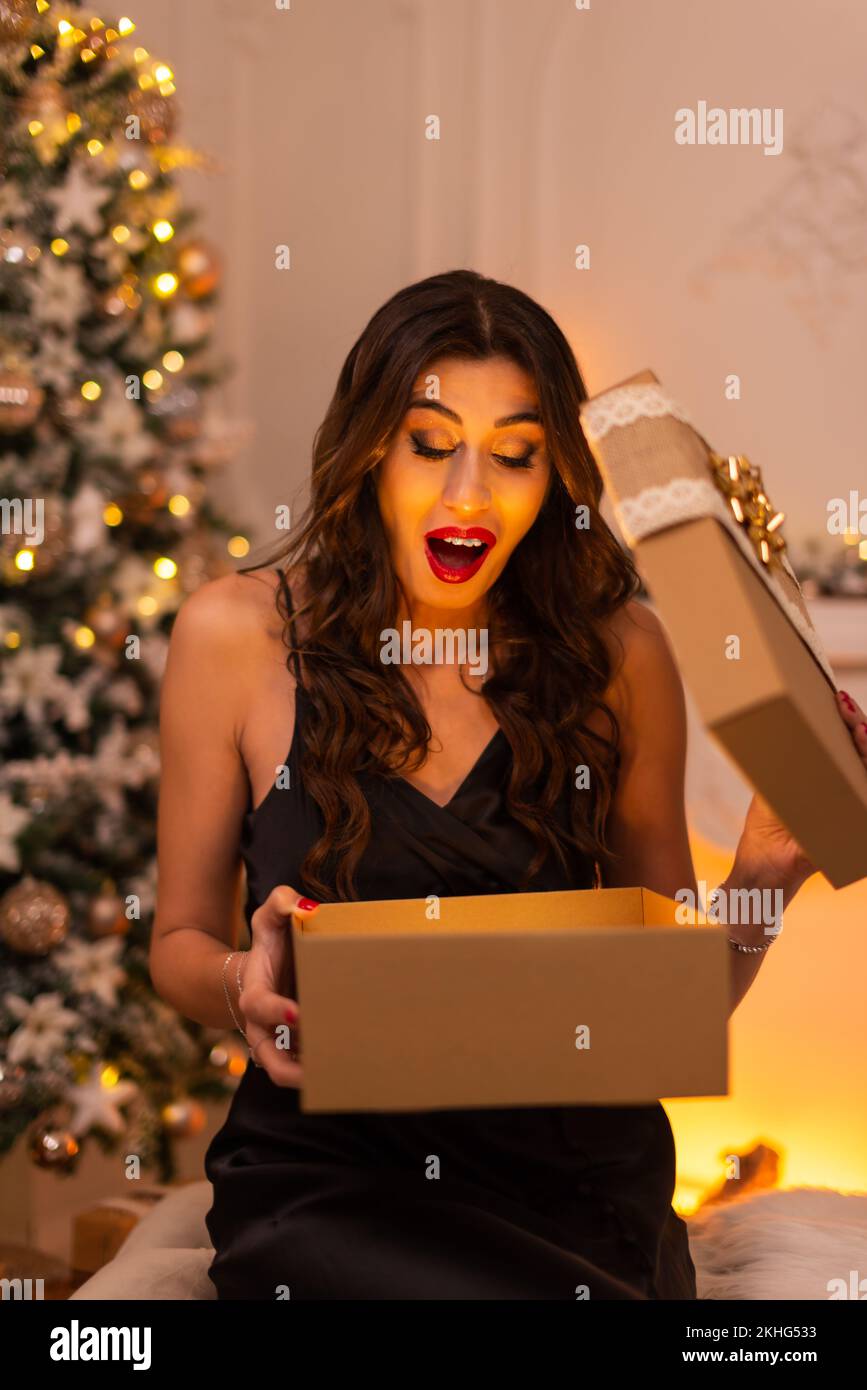 Portrait of a black dressed brunette woman with surprising emotion while opening a gift box there is a christmas tree and fireplace on the background. Stock Photo