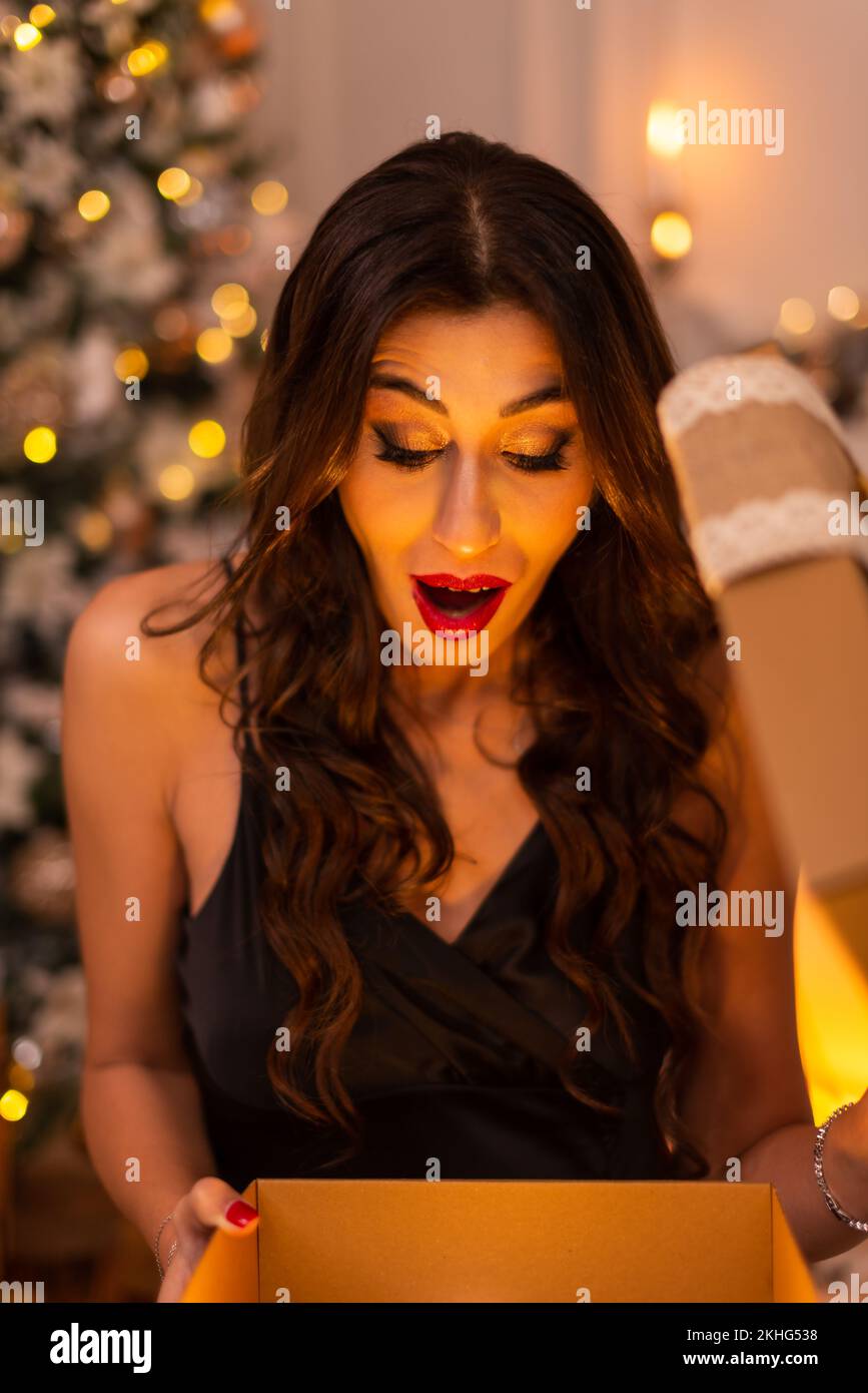 Portrait of a black dressed brunette woman with surprising emotion while opening a gift box there is a christmas tree and fireplace on the background. Stock Photo
