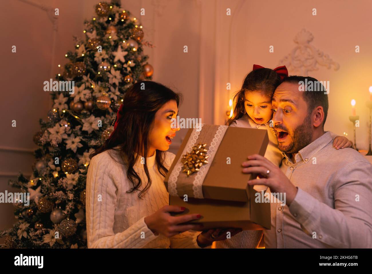 Happy family with surprising emotion while opening a gift box there is a christmas tree, fireplace and candles on the background. Stock Photo