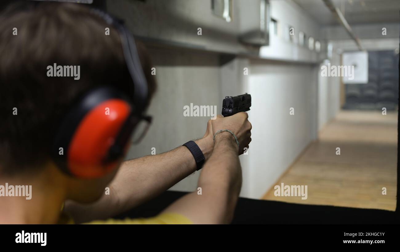 Man in tactical headphones aiming gun at target in shooting range Stock