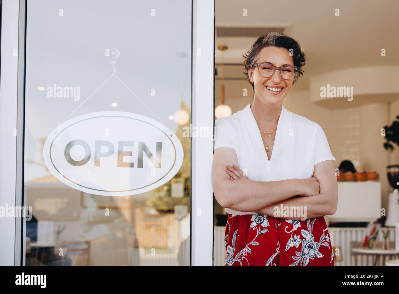 Happy cafe owner smiling at the camera while standing next to an open sign at the doorway of her coffee shop. Friendly small business owner welcoming Stock Photo