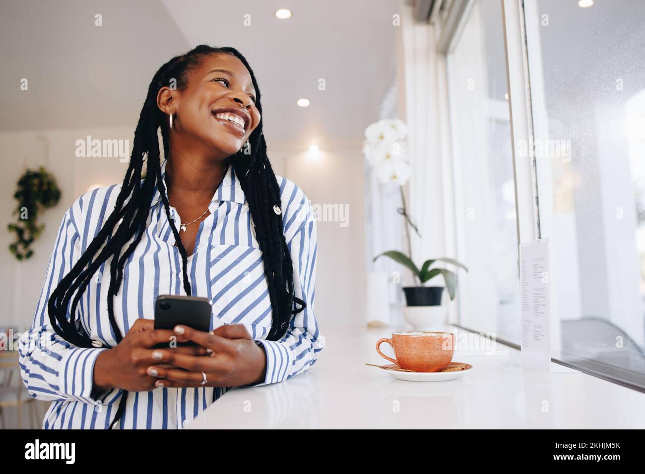 Young businesswoman smiling and looking away while holding a smartphone in a coffee shop. Happy young black businesswoman enjoying her coffee break in Stock Photo