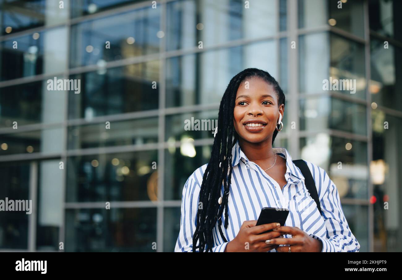 Pensive businesswoman holding a smartphone and wearing earphones during her work commute in the city. Young black business woman listening to music on Stock Photo