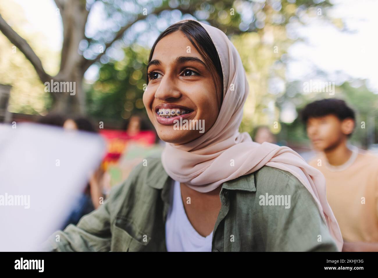 Muslim girl smiling happily while sitting with a group of demonstrators at a climate protest. Multicultural youth activists joining the global climate Stock Photo