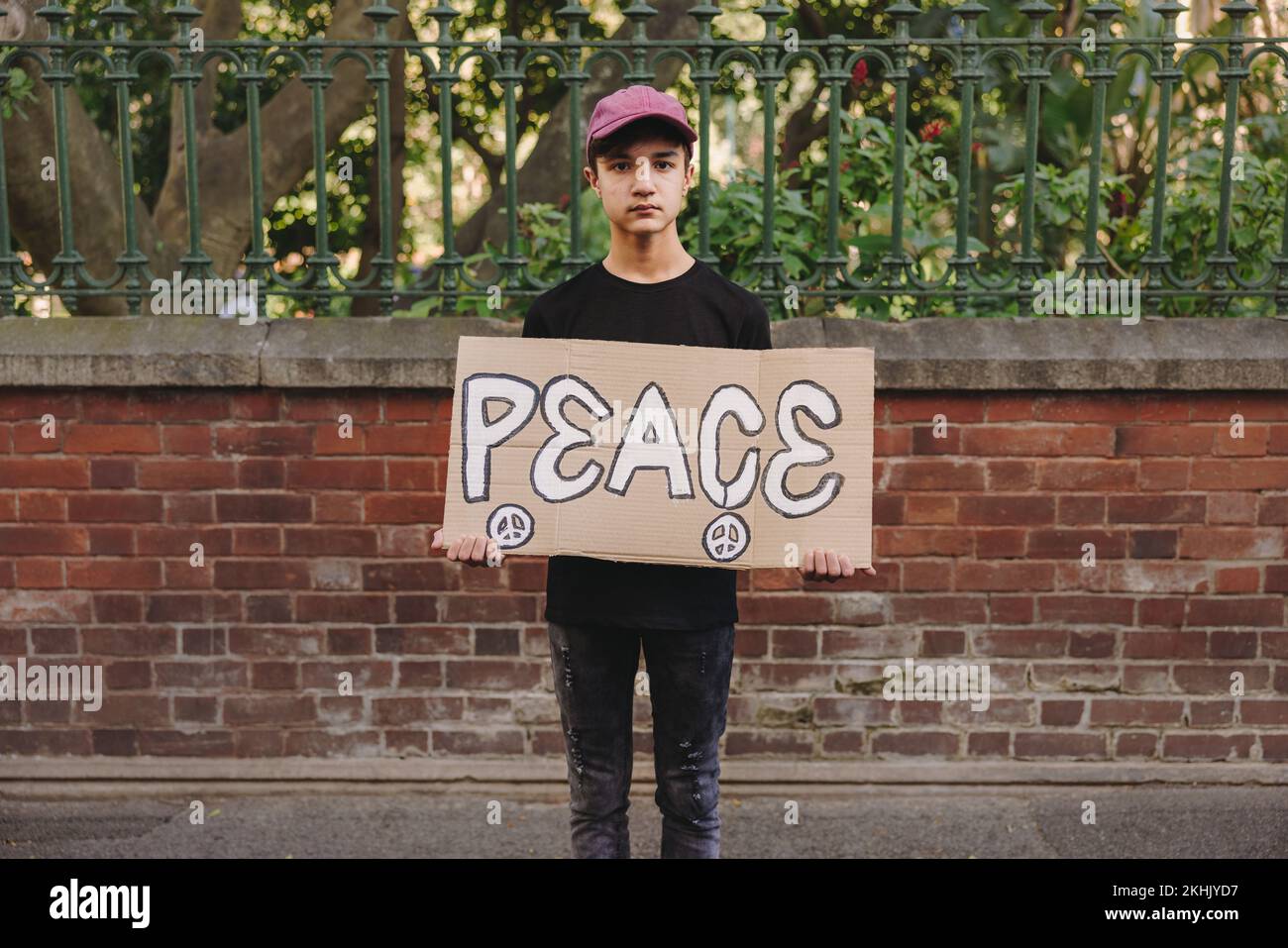 Young teenage boy looking at the camera while holding a peace poster. Young peace activist protesting against war and violence. Stock Photo