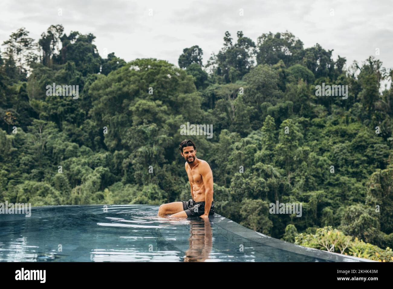 Happy young man sitting in swimming pool and looking away. Caucasian man enjoying his holiday at luxury resort in nature. Stock Photo