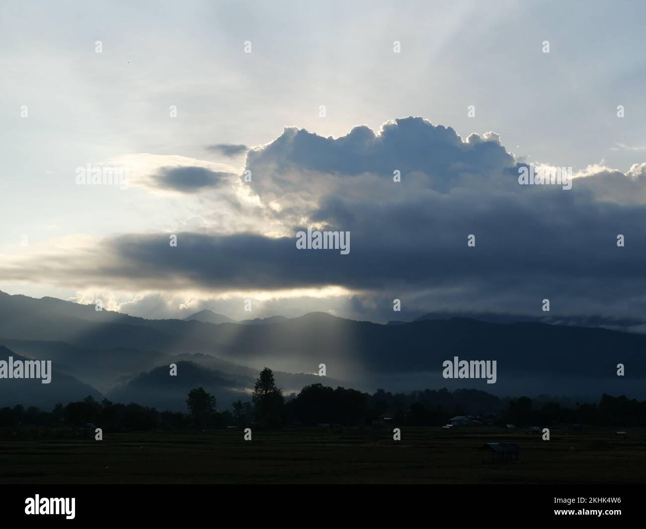 Silhouette of tree and mountain with sunbeam light shoot through the dark cloud to the land at sunrise, Mist covers the forest and mountains at dawn Stock Photo