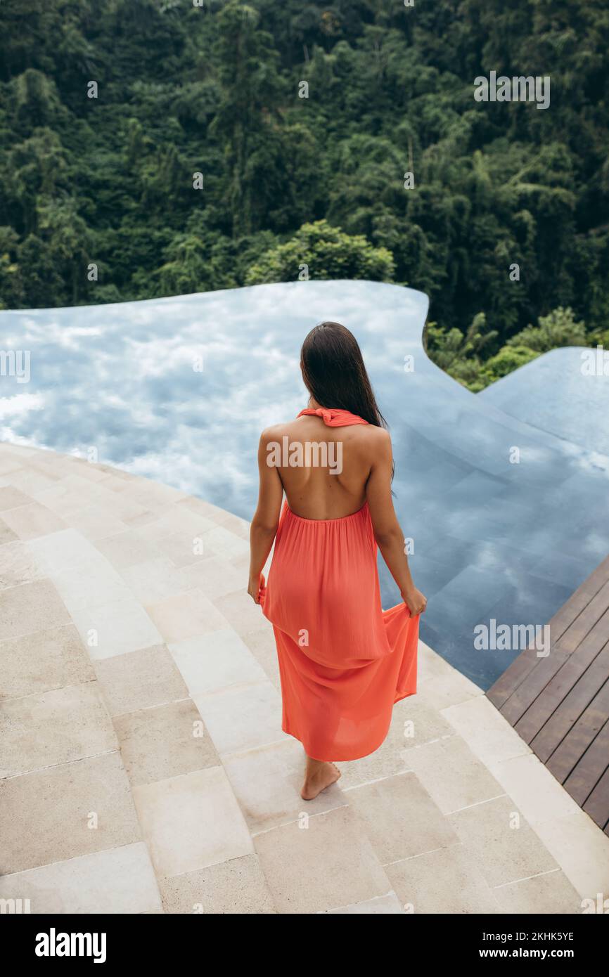 Rear view shot of young woman walking towards swimming pool at luxury resort. Female model in sundress at poolside. Stock Photo