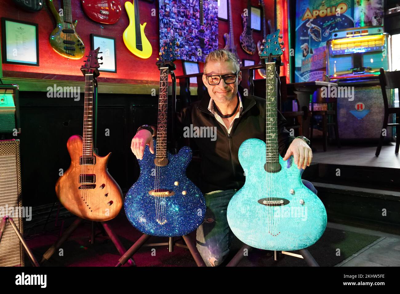 24 November 2022, Hamburg: Guitar maker Jens Ritter kneels behind his guitars "25th Anniversary of Magic & Freedom" (M), studded with more than 11,000 Swarovski stones, and the guitar "The Paraiba Velvet" (r) at the Zwick St. Pauli. Ritter presents some of his instruments in the cult pub "Zwick" in Hamburg-St. Pauli. His electric guitars and electric basses are purchased not only by musicians but also by collectors and museums; his instruments are also on display at the Metropolitan Museum of Art (New York) at the Smithsonian American Art Museum (Washington, DC) and at the Technoseum (Mannhe Stock Photo