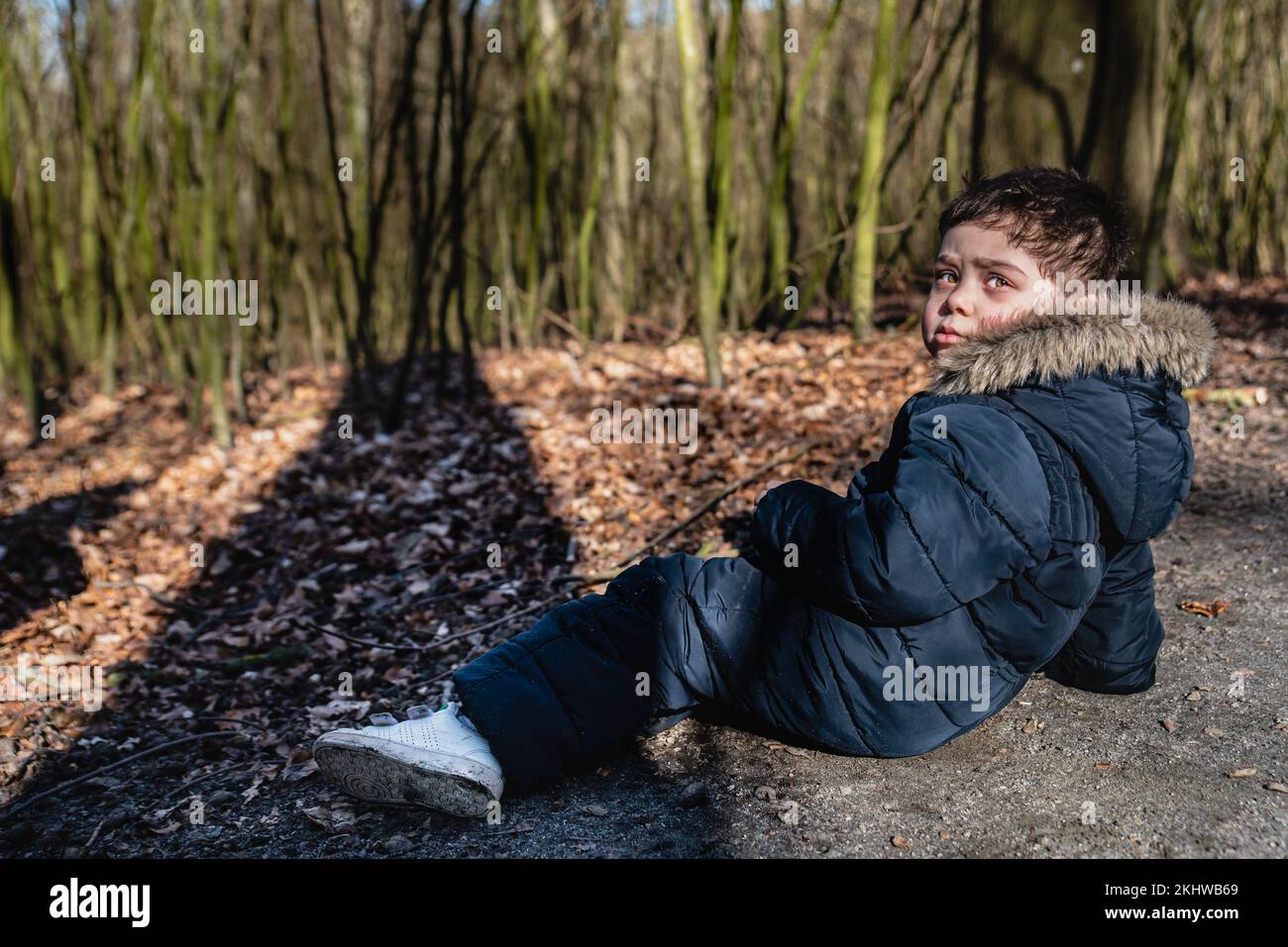 Kid with snowsuit lying on the ground in the woods. Serious child dressing snow suit is relaxing on the forest 's floor with face lit by the sunlight Stock Photo