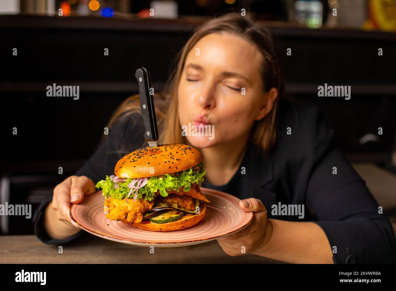 Joyful woman with close eyes presenting, kiss double hamburger with knife in bun on plate in cafe. Food delivery order Stock Photo