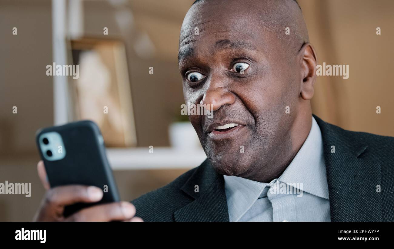 African American old 50s surprised amazed shocked man looking at mobile phone screen reading unbelievable news waving head no disagreement negation Stock Photo