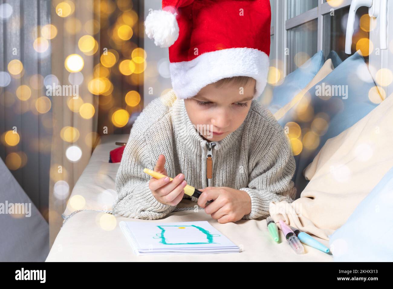 A wish list for a Christmas miracle. A serious focused kid in a Santa hat writes a wish list or a letter to Santa at home. The boy draws a phone. A ch Stock Photo