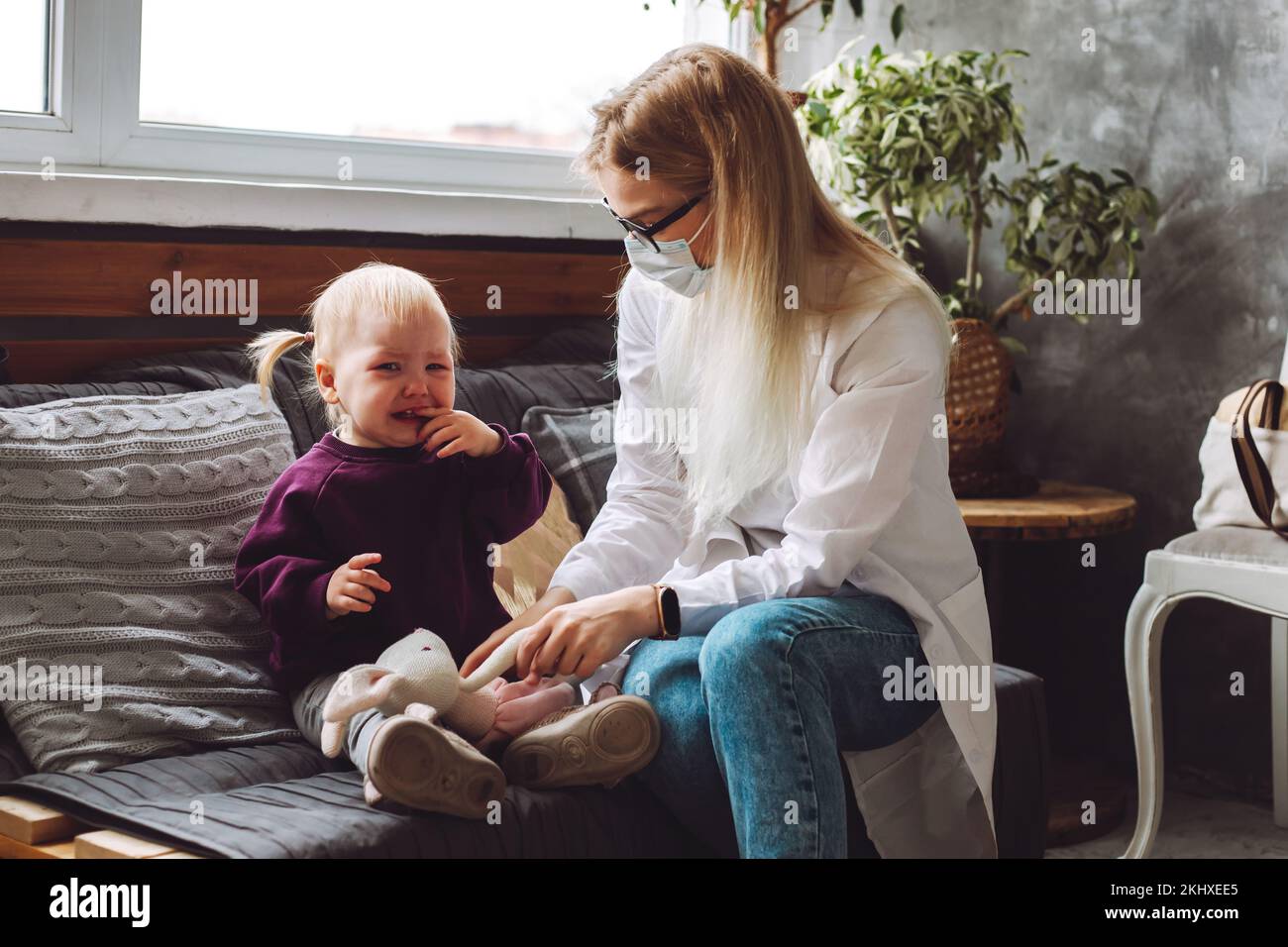 Portrait of sad little girl toddler sitting on sofa, crying near childrens doctor, holding soft toy rabbit at home. Stock Photo
