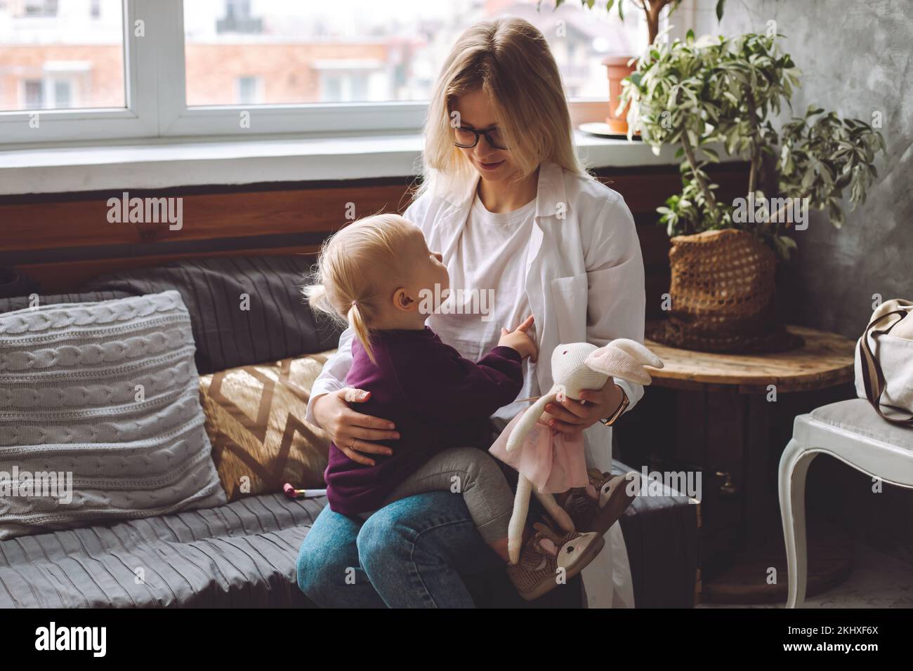 Side view of little blond girl sit on childrens doctor, looking at pediatrician visiting patient at home. Nurse visit. Stock Photo