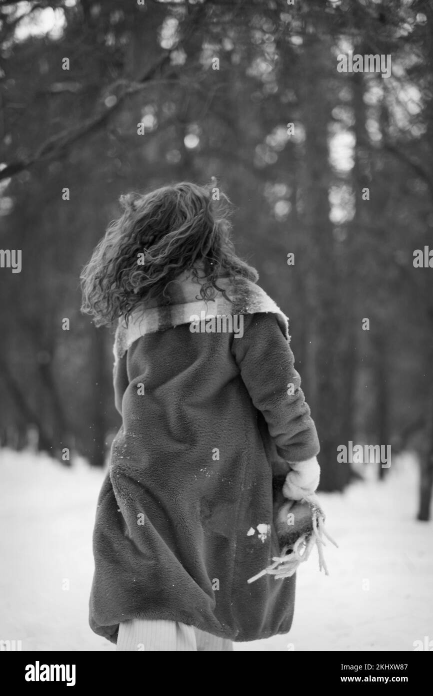 A white blonde girl in a knitted hat, a long scarf and a fur coat walks through the forest and smiles. Woman catches snow in white mittens. Winter is on the street. High quality photo. Christmas Eve in the park. A girl of European appearance enjoys life. New Year's mood, gifts, smiles, sale and joy Stock Photo