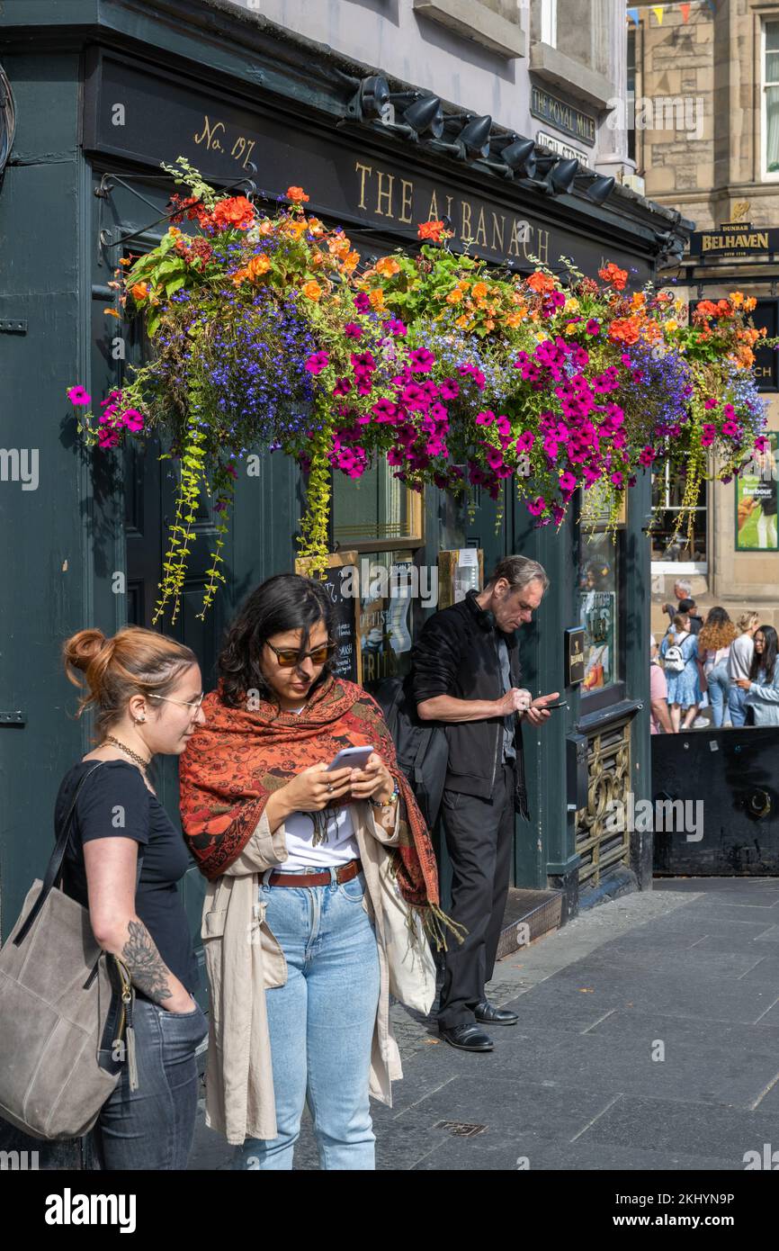 Tourists check their phones under the colourful hanging baskets of The Albanach, a traditional Scottish Bar and Restaurant on Edinburgh's Royal Mile. Stock Photo