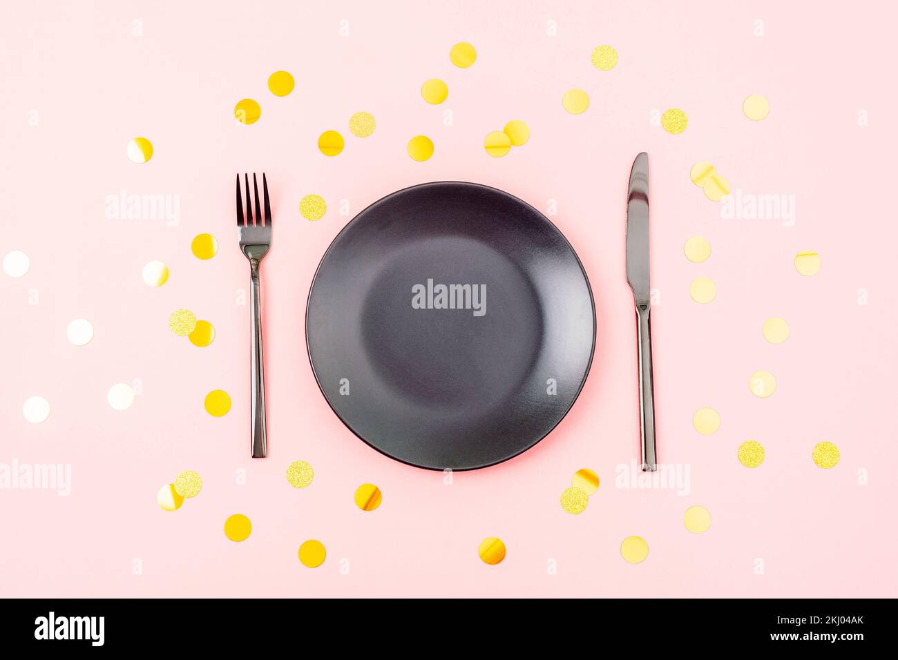 Stylish table setting with black plate, cutlery on pink background with golden confetti. Christmas, birthday, holiday concept. Top view, flat lay. Stock Photo
