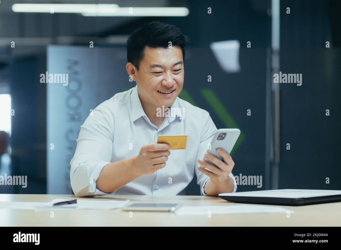 Young Asian man sitting at desk in office, paying with credit card online, making order by phone, making transaction with mobile banking app. Stock Photo