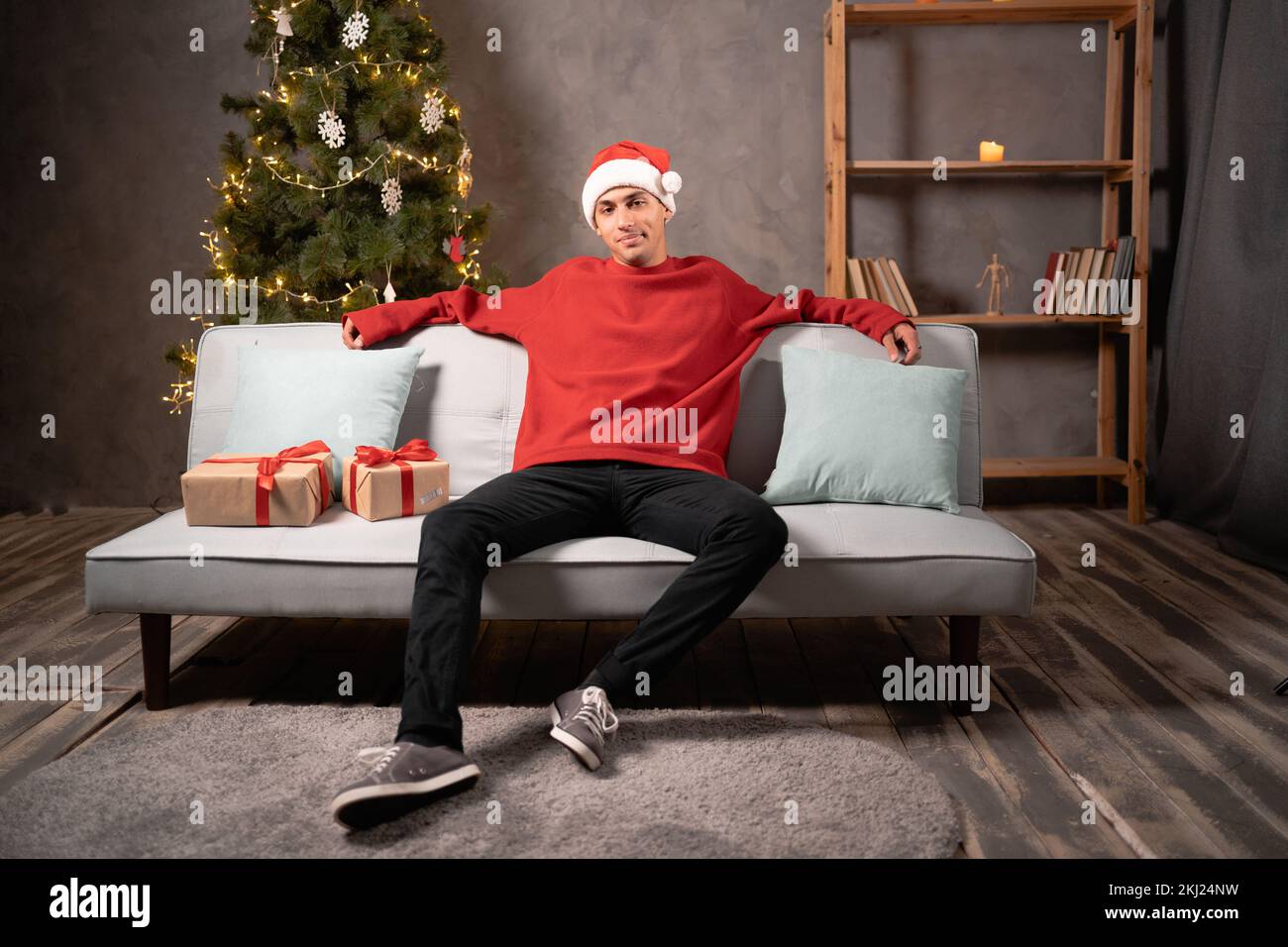 Relaxed young man in Santa hat sit rest on couch in Christmas living room daydreaming, calm male relax on sofa at home, relieve negative emotion, stre Stock Photo