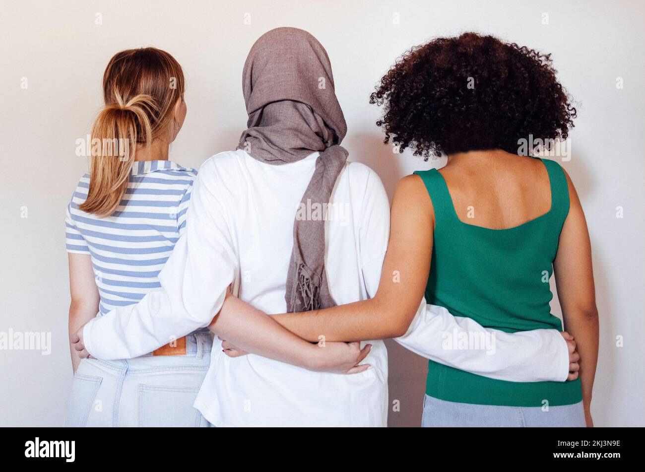 Strong female friendship. Rear view ttree teen girls best friends holding hands behind back and hugging while standing in front of beige wall outdoors Stock Photo