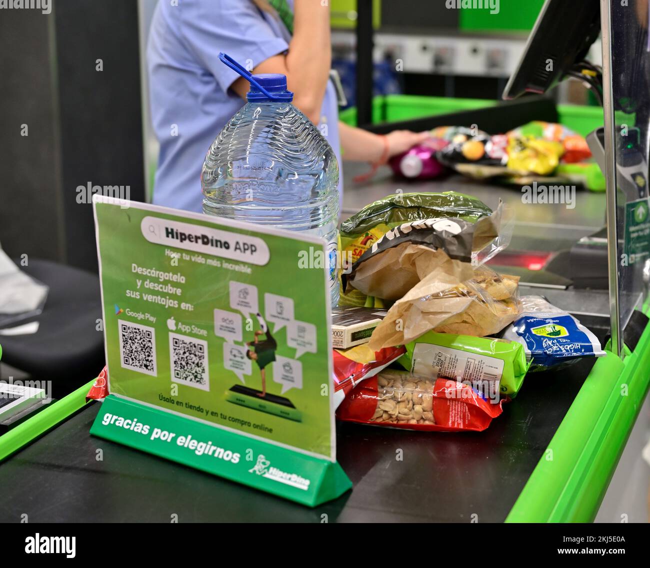 Check out counter at a HiperDino supermarket with food items on conveyor belt before staff at till Stock Photo